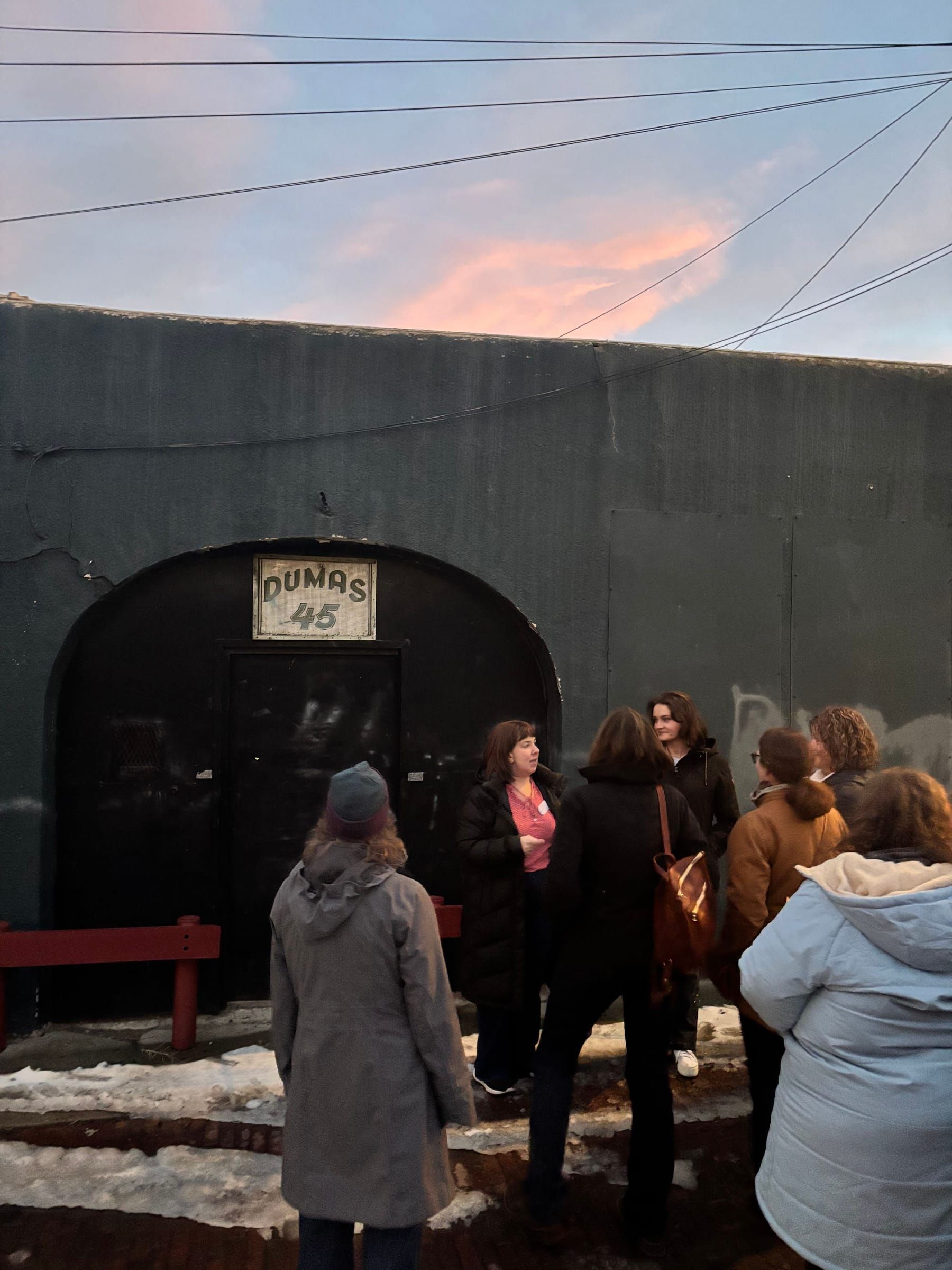 A tour group gathered at the arched DUMAS No.45 entrance at dusk, the guide telling the house's story under a pink Butte sky